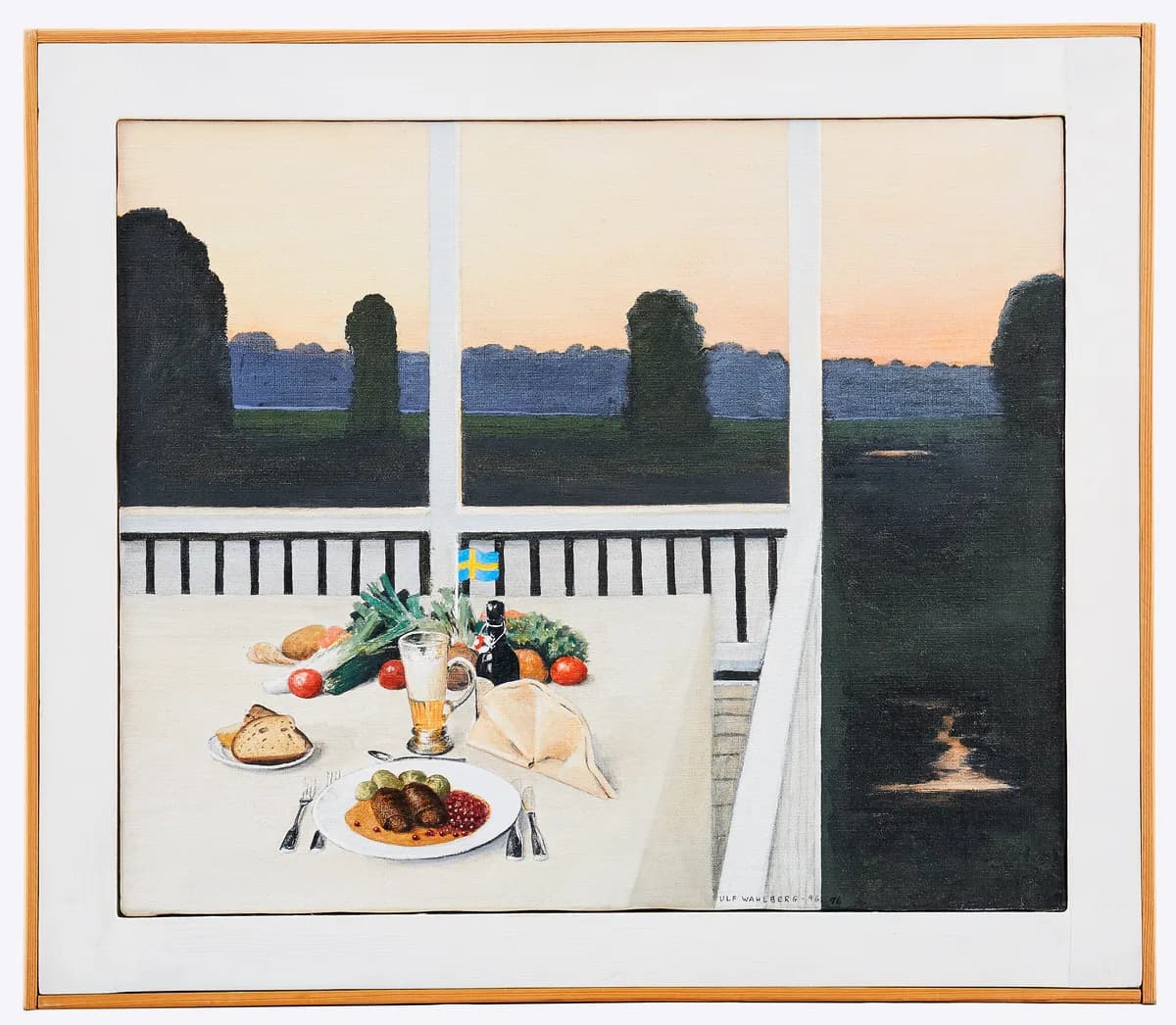 A set dinner table on a veranda at dusk, with a plate of meatballs, bread, vegetables, and a small Swedish flag, with a lake and forest visible in the background.